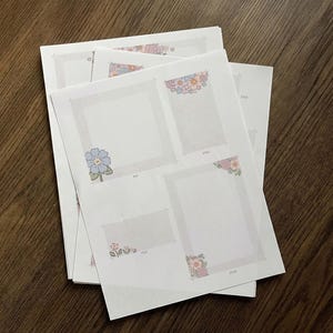 Peut inclure: Une pile de feuilles de papier blanches avec des motifs floraux dans les coins et des contours de cadres. Les motifs floraux comprennent des fleurs bleues, roses et oranges. Les feuilles sont sur une surface en bois brun foncé.