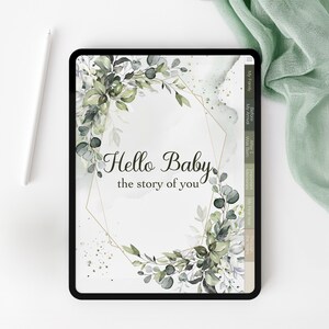 以下が含まれることがあります： 黒いタブレットには「Hello Baby, the story of you」というテキストが表示され、花のリースで囲まれています。白いスタイラスと緑色の布がタブレットの隣にあります。