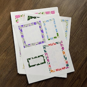 May include: A stack of white paper sheets featuring floral frame designs in various colors. The frames include purple, blue, green, and orange flowers and foliage. The paper has a textured background.