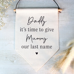 May include: A cream-coloured banner with the words "Daddy it's time to give Mummy our last name" in black script. A small black heart is at the bottom. The banner is suspended from a wooden dowel with a brown cord.