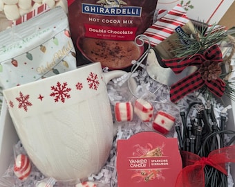 May include: A gift box filled with a white snowflake-decorated mug, Ghirardelli hot cocoa mix, marshmallows, peppermint candies, a Yankee Candle, and a decorative sled. The box includes a card that reads "SIT LOOK DRINK".