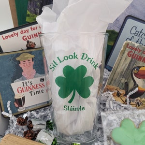 Puede incluir: Una caja de regalo llena de art&iacute;culos de tem&aacute;tica irlandesa, que incluyen un vaso con un tr&eacute;bol y el texto "Sit Look Drink Sl&aacute;inte", un letrero de madera con el texto "SIT. LOOK. DRINK." y una galleta de tr&eacute;bol verde.
