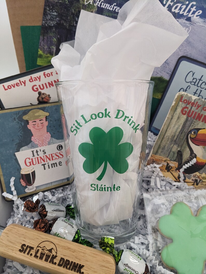 Puede incluir: Una caja de regalo llena de art&iacute;culos de tem&aacute;tica irlandesa, que incluyen un vaso con un tr&eacute;bol y el texto "Sit Look Drink Sl&aacute;inte", un letrero de madera con el texto "SIT. LOOK. DRINK." y una galleta de tr&eacute;bol verde.