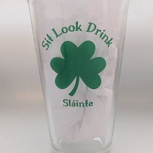 Puede incluir: Un vaso de pinta de vidrio transparente con un tr&eacute;bol verde y el texto "Sit Look Drink" y "Sl&aacute;inte" impreso en &eacute;l.