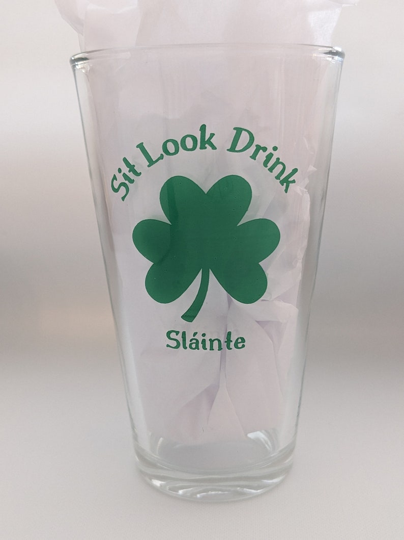 Puede incluir: Un vaso de pinta de vidrio transparente con un tr&eacute;bol verde y el texto "Sit Look Drink" y "Sl&aacute;inte" impreso en &eacute;l.