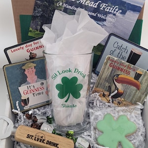Puede incluir: Una caja de regalo verde y blanca llena de art&iacute;culos de tem&aacute;tica irlandesa, que incluyen un vaso con el texto "Sit Look Drink" y un dise&ntilde;o de tr&eacute;bol, un abridor de botellas con el mismo texto, una galleta de tr&eacute;bol verde y varios posavasos con la marca Guinness. La caja tambi&eacute;n incluye una tarjeta con el texto "Cead Mead Failte (A Hundred Thousand Welcomes)".