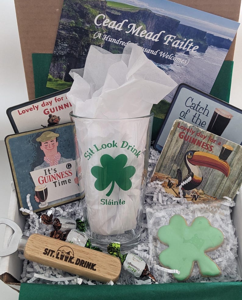 Puede incluir: Una caja de regalo verde y blanca llena de art&iacute;culos de tem&aacute;tica irlandesa, que incluyen un vaso con el texto "Sit Look Drink" y un dise&ntilde;o de tr&eacute;bol, un abridor de botellas con el mismo texto, una galleta de tr&eacute;bol verde y varios posavasos con la marca Guinness. La caja tambi&eacute;n incluye una tarjeta con el texto "Cead Mead Failte (A Hundred Thousand Welcomes)".