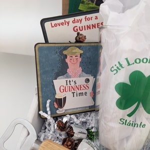 Puede incluir: Una caja de regalo llena de art&iacute;culos para un amante de la Guinness. La caja incluye un vaso con un tr&eacute;bol y el texto "Sit Look", un abridor de botellas con mango de madera, una posavasos con un anuncio vintage de Guinness y caramelos de chocolate.