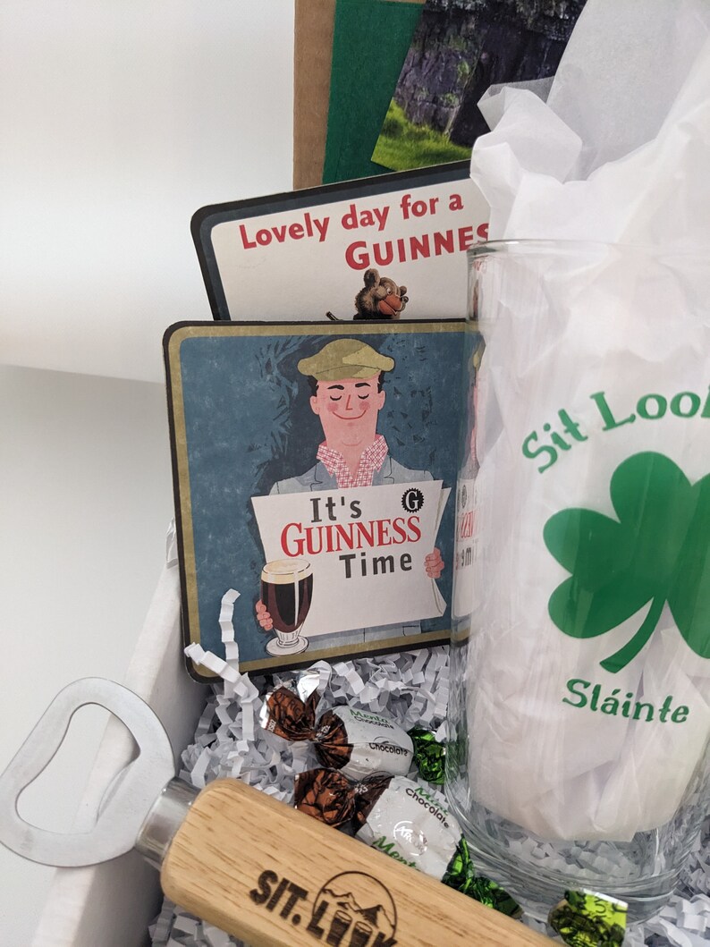 Puede incluir: Una caja de regalo llena de art&iacute;culos para un amante de la Guinness. La caja incluye un vaso con un tr&eacute;bol y el texto "Sit Look", un abridor de botellas con mango de madera, una posavasos con un anuncio vintage de Guinness y caramelos de chocolate.