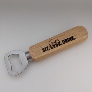 Puede incluir: Un abridor de botellas de madera con un abridor de metal y un mango de madera. El mango tiene el texto "SIT. LOOK. DRINK." grabado.