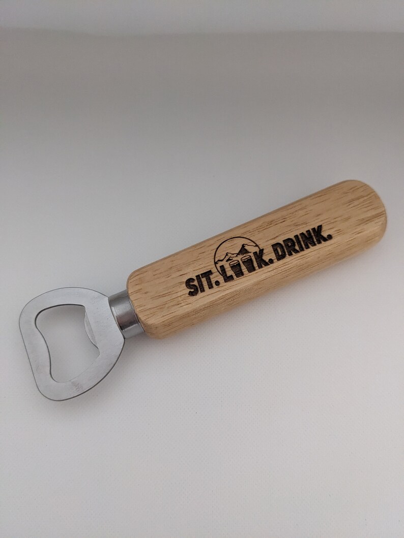 Puede incluir: Un abridor de botellas de madera con un abridor de metal y un mango de madera. El mango tiene el texto "SIT. LOOK. DRINK." grabado.