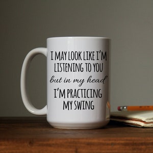 May include: White ceramic coffee mug with black text that reads "I may look like I'm listening to you but in my head I'm practicing my swing."