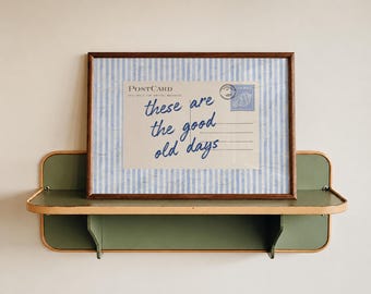 May include: A framed print featuring a postcard design with light blue and white vertical stripes. The postcard text reads "POSTCARD" and "these are the good old days" in blue script, with a blue stamp graphic in the upper right corner.