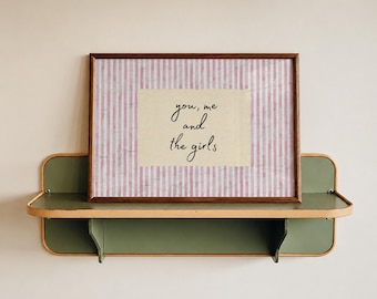 May include: A rectangular framed print featuring a light pink and white vertical striped background with a central cream-colored rectangle containing the black cursive text you, me and the girls, resting on a green and light wood wall shelf.