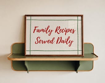 May include: A framed sign with the text "Family Recipes Served Daily" in red script. The sign is set against a cream-colored background with a green border. It rests on a green and tan shelf, mounted on a neutral wall.