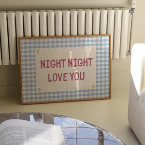 May include: A framed wall art piece with a light blue and white gingham pattern border. The center features a linen-colored rectangle with the words "NIGHT NIGHT LOVE YOU" in pink lettering. The frame is a light brown color.