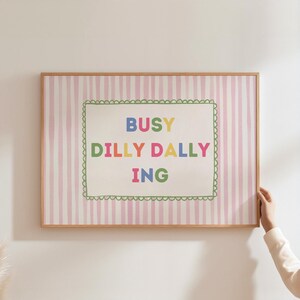 May include: A framed print with a pink and white striped background. The print features the words "BUSY DILLY DALLY ING" in colorful letters within a green scalloped border. The frame is a light brown color.