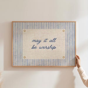 May include: A framed artwork with a light blue and white striped background. The center features a beige rectangle with the words "may it all be worship" in navy blue script. The frame is a light brown color.
