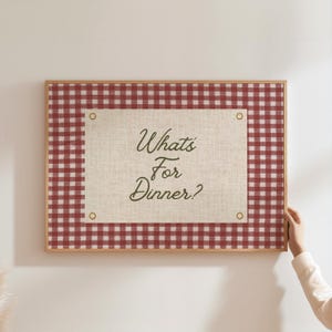 May include: A framed decorative board with a red and white checkered pattern border. The center features a beige linen-like material with the text "What's For Dinner?" in a script font. The frame is a light wood color.