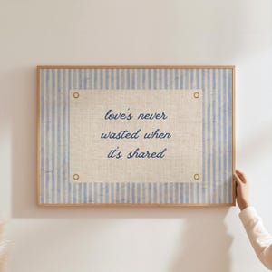 Puede incluir: Una obra de arte enmarcada con un fondo a rayas azul claro y blanco. La obra presenta un panel rectangular central con la cita "love's never wasted when it's shared" en escritura azul marino. El marco es de madera marrón claro.
