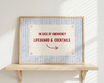 May include: A framed print with a light wood frame. The print has a blue and white striped border and a central off-white rectangle with the text "IN CASE OF EMERGENCY LIFEGUARD & COCKTAILS" in red. A red arrow points to the right.