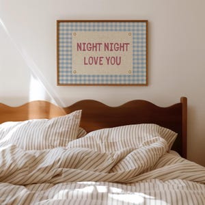 May include: A framed wall art piece with the text "NIGHT NIGHT LOVE YOU" in a pink font. The artwork has a light blue gingham pattern border and a natural linen-colored center. The bed has striped bedding and a wooden headboard.