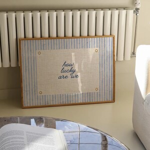 May include: A framed artwork with a light blue and white striped border and a beige center. The artwork features the text "how lucky are we" in a handwritten style. The frame is a light brown color.