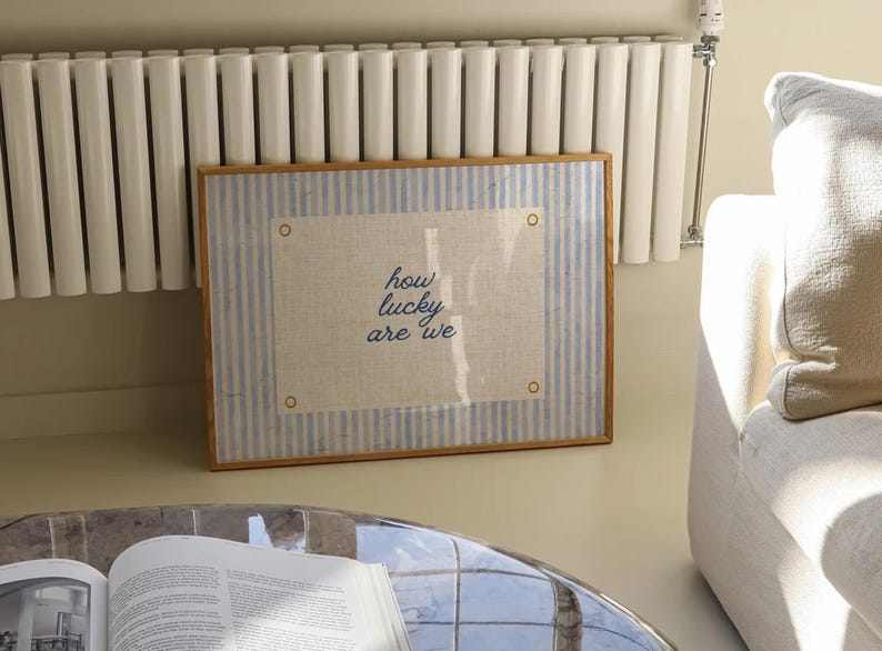 May include: A framed artwork with a light blue and white striped border and a beige center. The artwork features the text "how lucky are we" in a handwritten style. The frame is a light brown color.