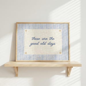以下が含まれることがあります： 「these are the good old days」のテキストが入った額入りのプリント。プリントには、水色と白のストライプのボーダーと、中央にオフホワイトの長方形があります。フレームは明るい木の色です。プリントは木製の棚に飾られています。
