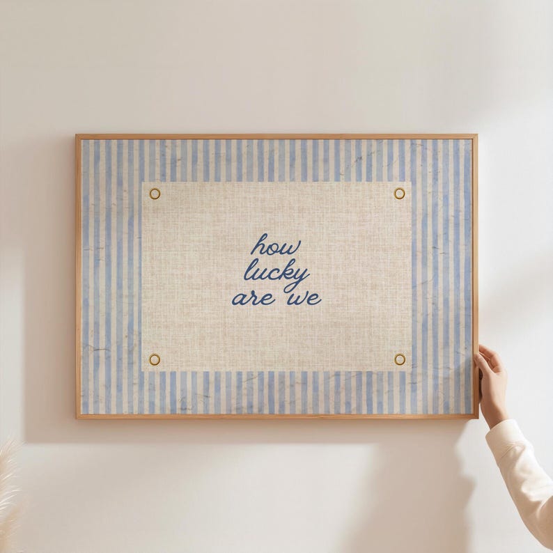 May include: A framed artwork with a light blue and white striped background and a central linen-textured panel. The panel features the handwritten text "how lucky are we" in dark blue script. The frame is made of light-colored wood.