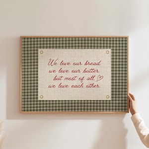 Op de afbeelding: Een ingelijst wandkunstwerk met een groen-witte geruite rand. Het middenstuk is een canvas met de tekst "We love our bread, we love our butter, but most of all, we love each other" in rood schrift.