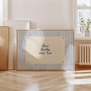 May include: Framed artwork with a light blue and white striped background. The artwork features a central rectangular panel with the words "how lucky are we" in a cursive font. The frame is a light wood color.