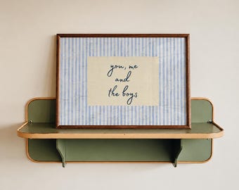 May include: A rectangular framed print featuring a light blue and white vertical striped background with a central cream-colored rectangle containing the dark blue handwritten text "you, me and the boys" displayed in a simple wooden frame.