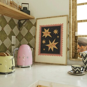 May include: A pink electric kettle with a silver handle and base sits on a white countertop next to a yellow toaster. A framed print with a black background and pink and yellow stars with faces hangs on the wall behind the appliances.
