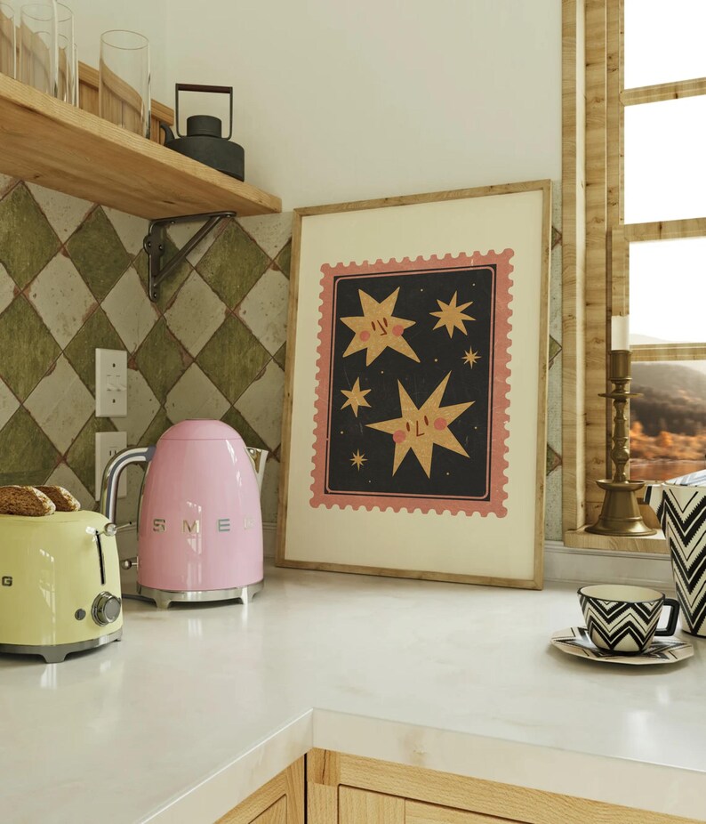 May include: A pink electric kettle with a silver handle and base sits on a white countertop next to a yellow toaster. A framed print with a black background and pink and yellow stars with faces hangs on the wall behind the appliances.