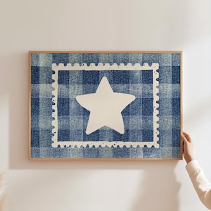 May include: Framed artwork with a white star inside a white postage stamp border, set against a blue and white plaid background. The artwork is in a light brown wooden frame, creating a rustic aesthetic.