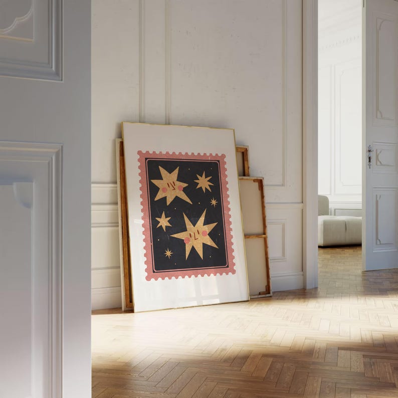 May include: A framed print with a pink and black design featuring three stars with faces. The stars are on a dark blue background with a pink border. The print is leaning against a white wall.