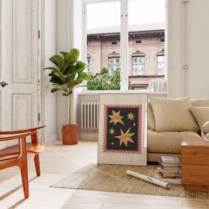 May include: A framed print with a black background and pink border features three yellow stars with faces. The stars are surrounded by smaller black stars. The print is hanging on a white wall in a living room setting.
