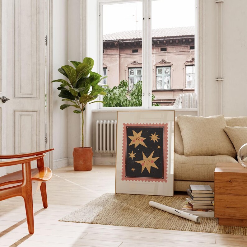 May include: A framed print with a black background and pink border features three yellow stars with faces. The stars are surrounded by smaller black stars. The print is hanging on a white wall in a living room setting.
