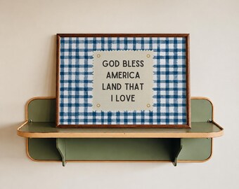 May include: A rectangular framed print featuring a blue and white gingham pattern background. Centered on the print is a cream-colored rectangular tag with the text "GOD BLESS AMERICA LAND THAT I LOVE" in black, accented by four small gold grommets.