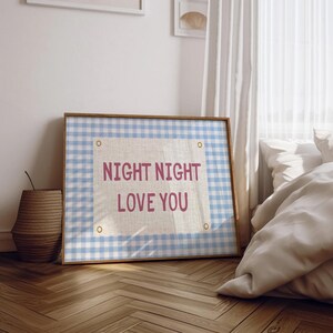 May include: A framed artwork with a light blue and white gingham pattern background. The artwork features the text "NIGHT NIGHT LOVE YOU" in pink. The frame is a light brown color. The artwork is displayed in a bedroom setting.