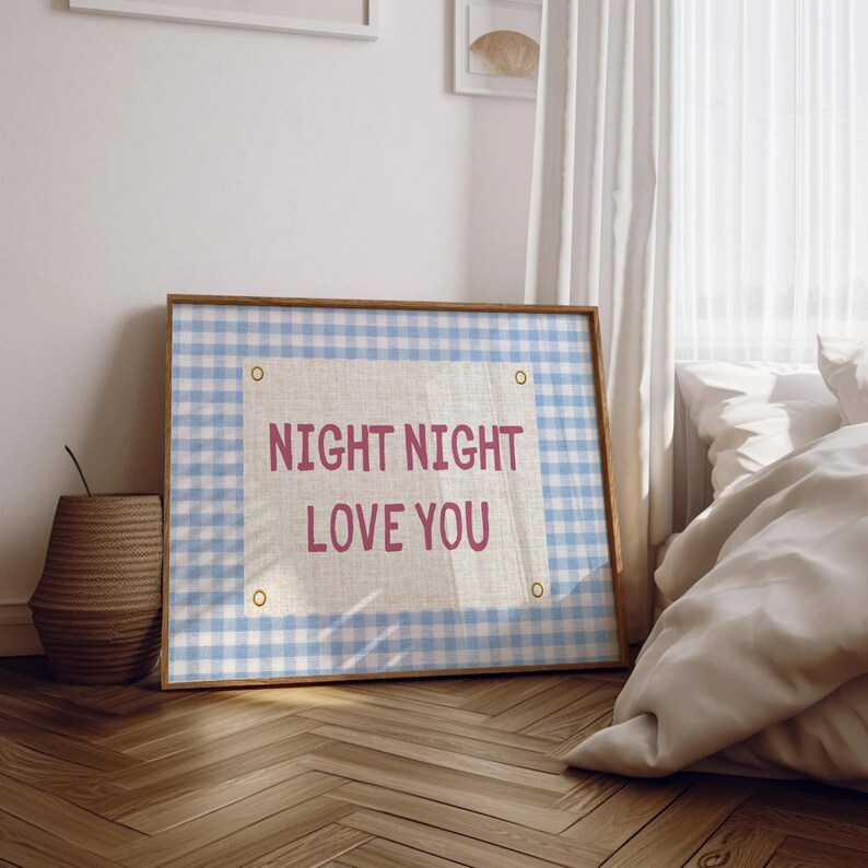 May include: A framed artwork with a light blue and white gingham pattern background. The artwork features the text "NIGHT NIGHT LOVE YOU" in pink. The frame is a light brown color. The artwork is displayed in a bedroom setting.