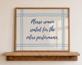 May include: A framed print with the text "Please remain seated for the entire performance." The print has a light beige background with a blue grid design. The frame is made of wood and sits on a wooden shelf.