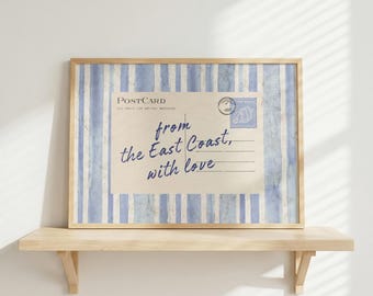 May include: A framed print featuring a vintage postcard design. The postcard has a light beige background with blue text that reads "from the East Coast, with love." The background has blue and white vertical stripes. The frame is made of light wood.