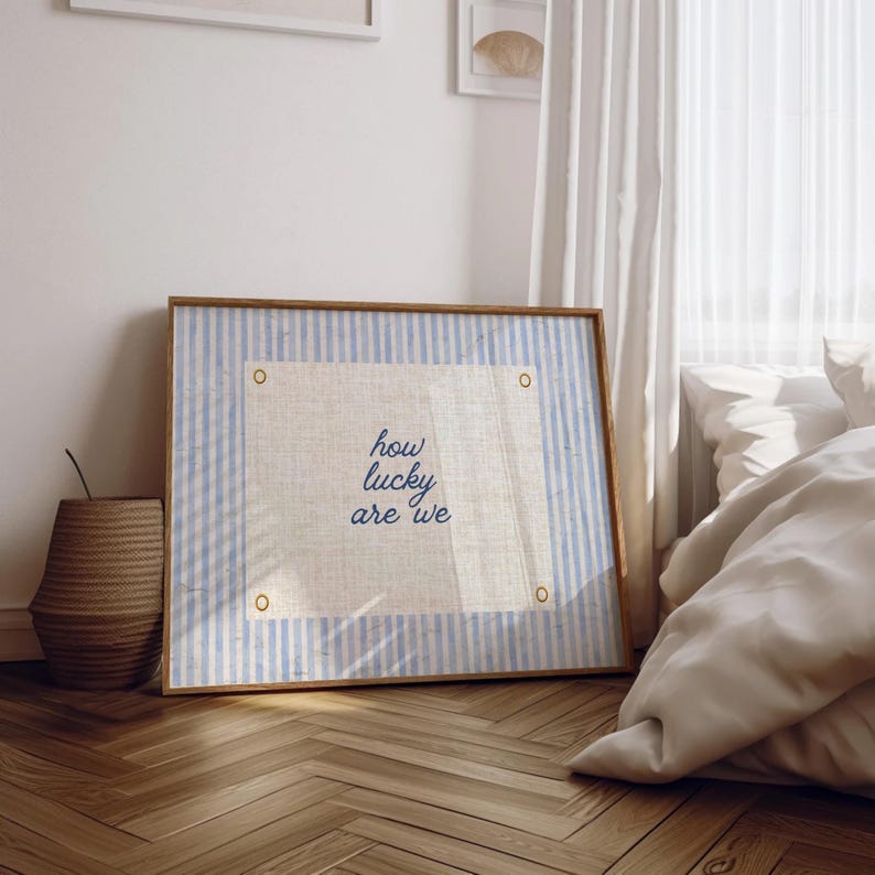 May include: A framed artwork with a light blue and white striped border and a central linen panel. The text "how lucky are we" is written in blue script. The frame is a light brown color. A woven basket is visible in the background.