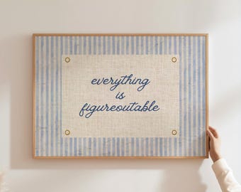 May include: A framed wall art piece with a light blue and white striped background. The center features a beige rectangle with the cursive text "everything is figureoutable" in dark blue. The frame is a light brown color.