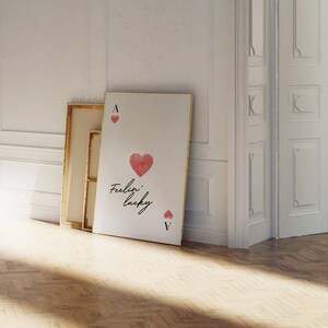 Op de afbeelding: Een witte canvas print met een gouden lijst met een rood hart met de tekst "Feelin' lucky" en de aas en vijf van harten speelkaarten.
