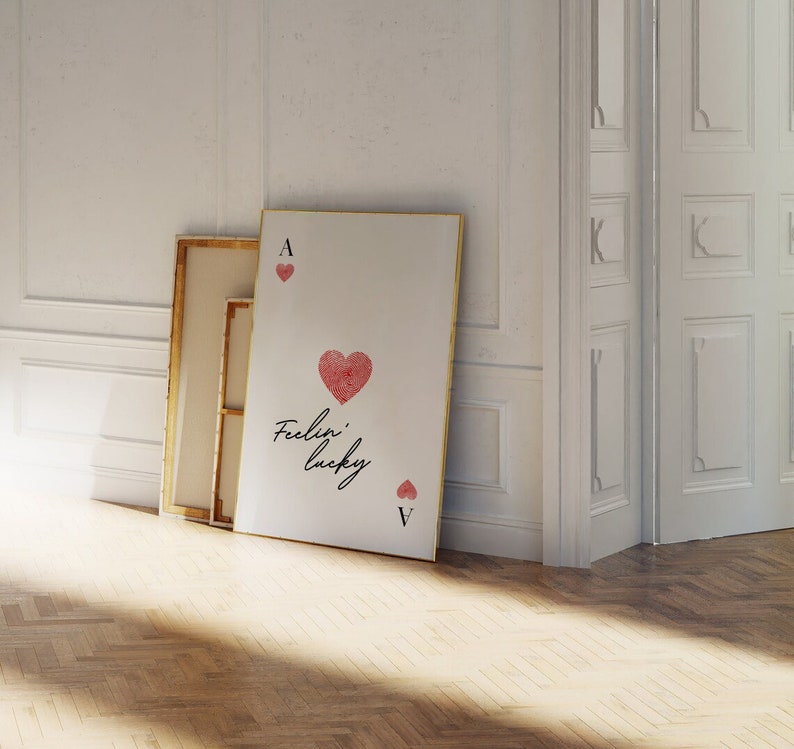 Op de afbeelding: Een witte canvas print met een gouden lijst met een rood hart met de tekst "Feelin' lucky" en de aas en vijf van harten speelkaarten.