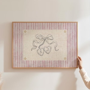 Puede incluir: Una obra de arte enmarcada con un fondo a rayas rosas y blancas. El diseño central es un boceto de un lazo, una cinta y dos colgantes en forma de corazón con las letras "K" y "L". El marco es de color marrón claro.