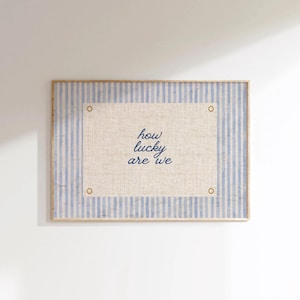 May include: A framed wall art piece with a light blue and white striped background. The center features a beige rectangle with the text "how lucky are we" in blue script. Gold-colored rivets are in each corner.